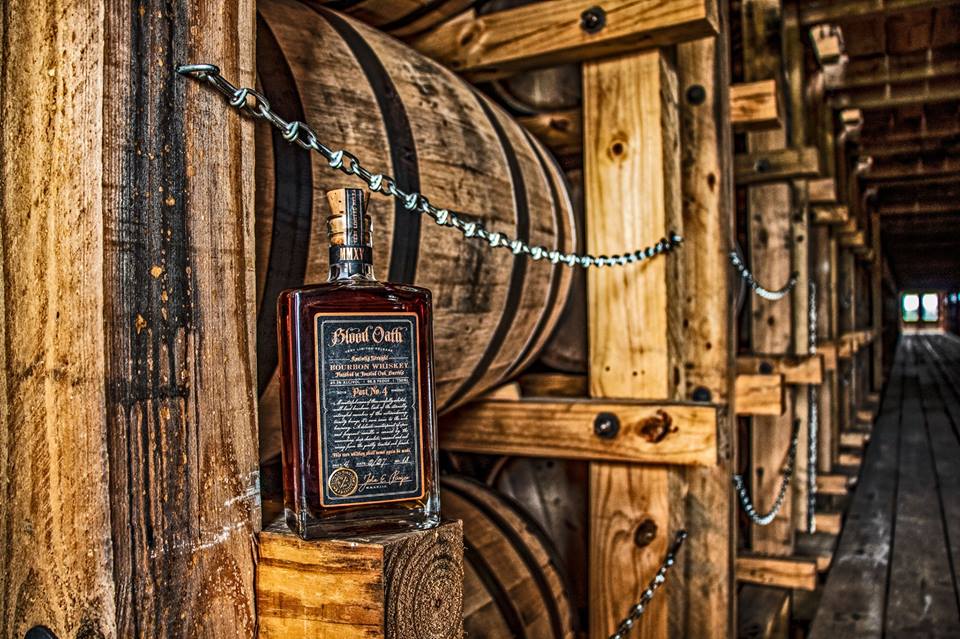bottle of blood oath in front of a couple bourbon barrels